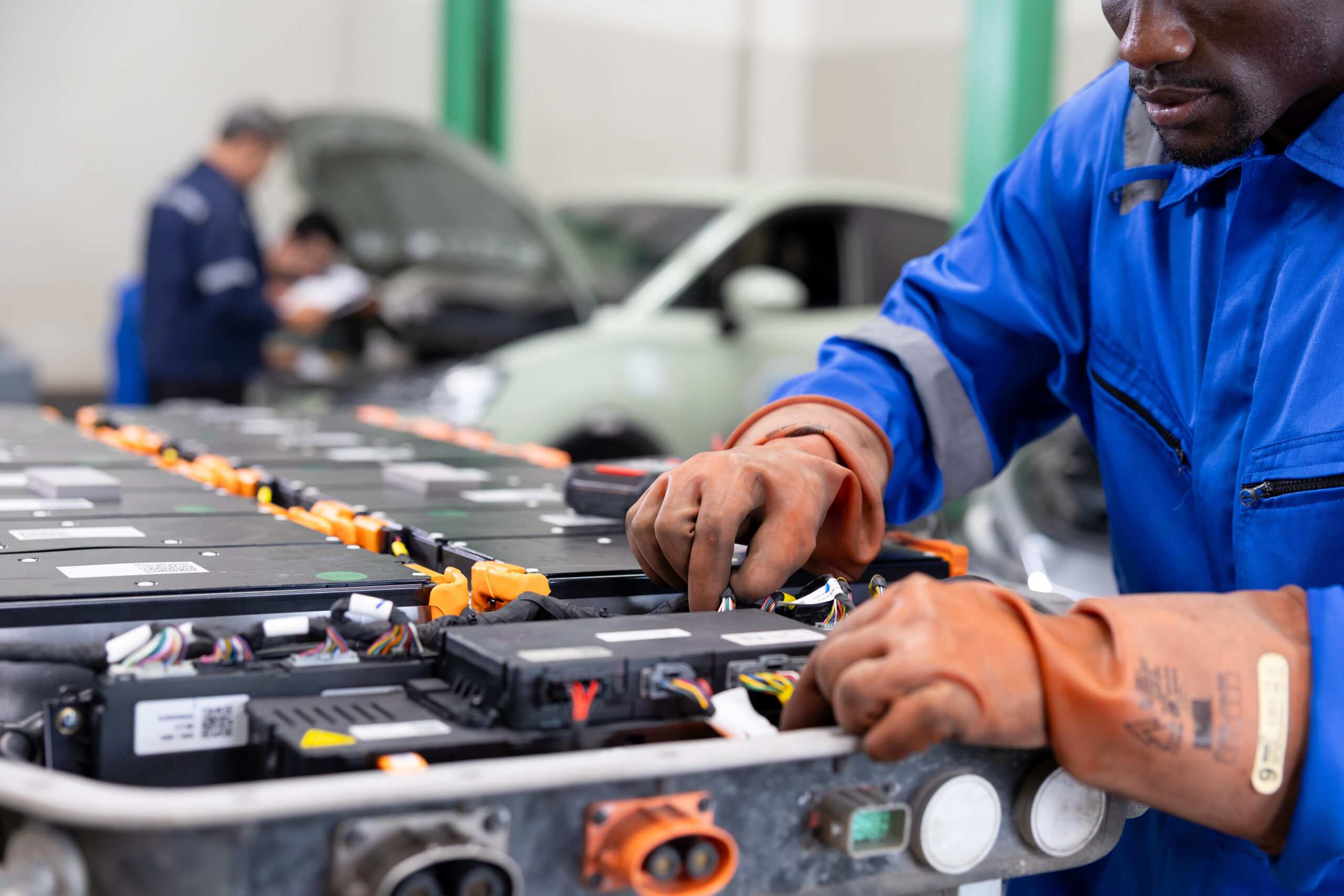 Technician wearing protective gloves services an electric vehicle battery pack in an auto workshop, inspecting modules and connections with tools beside a car.